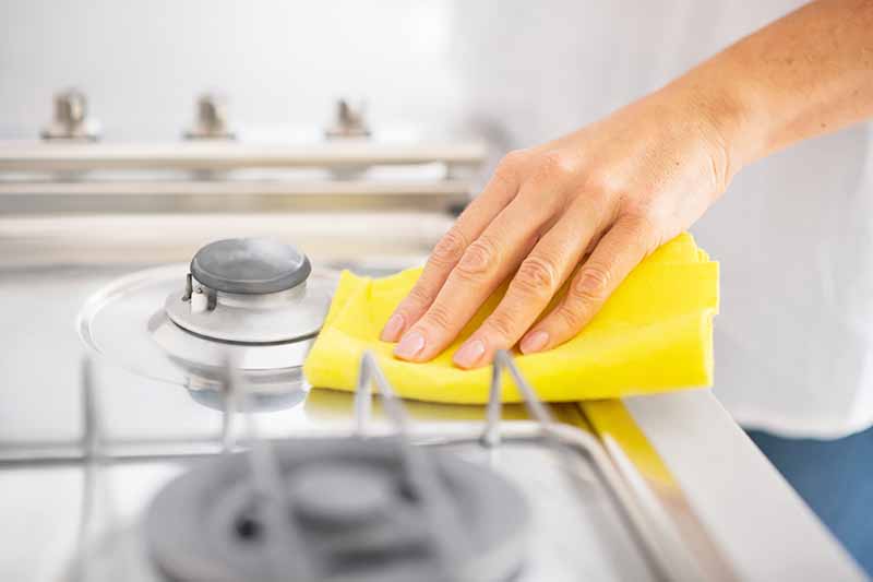 Horizontal image of a hand cleaning the stovetop of an oven with a yellow towel.