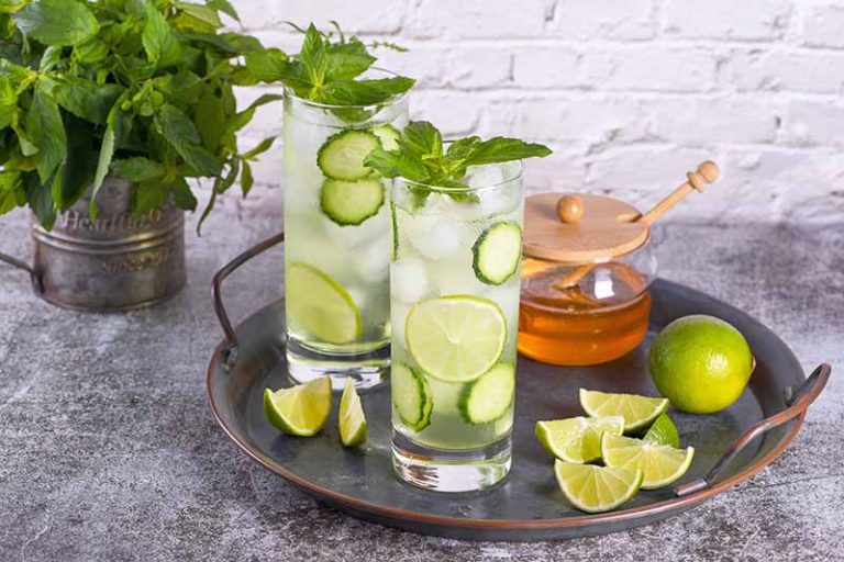 Horizontal image of two cocktails with fresh garnishes on a tray with limes and honey.