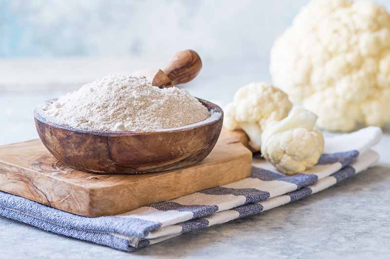 Horizontal image of a wooden bowl filled with cauliflower flour next to a whole head of fresh cauliflower on top of a striped napkin.