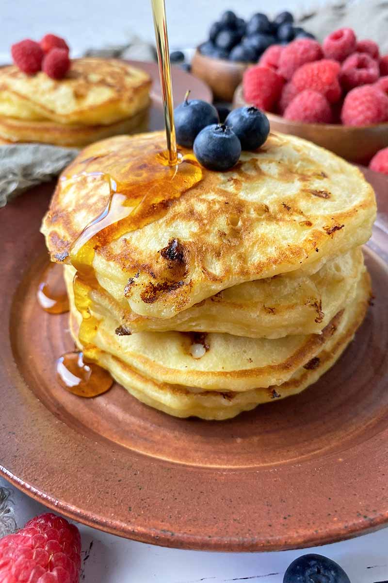 Vertical image of pouring syrup over a stack of pancakes topped with blueberries on a brown plate.