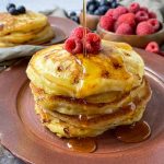 Horizontal image of a stack of pancakes with raspberries with maple syrup poured over the top on a brown plate in front of bowls of berries.