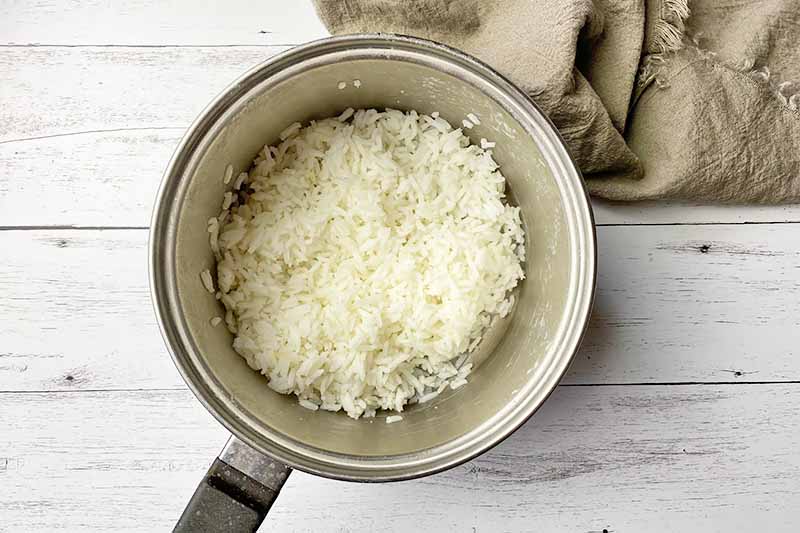 Horizontal image of fluffy cooked white grains in a small pot next to a tan napkin.