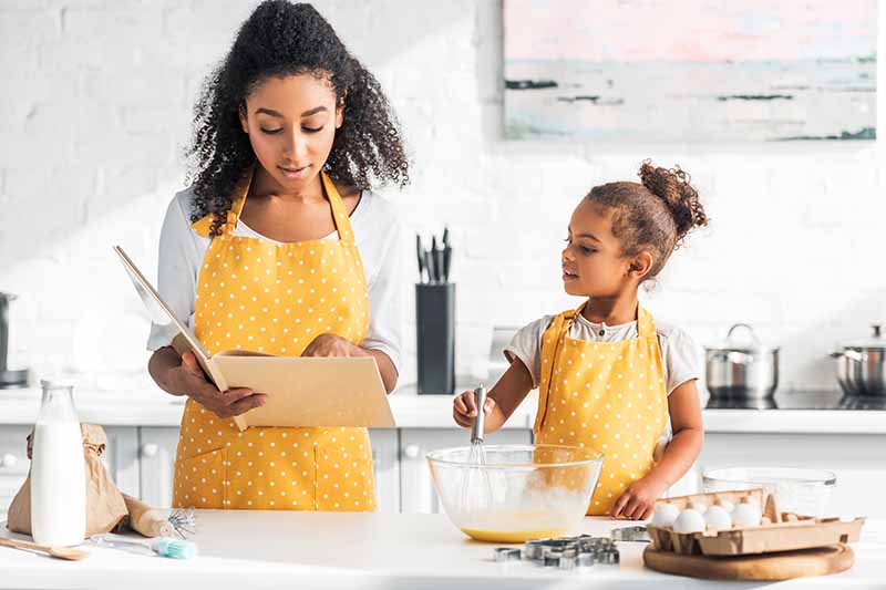 Horizontal image of a mom and daughter reading a cookbook while in the kitchen together.