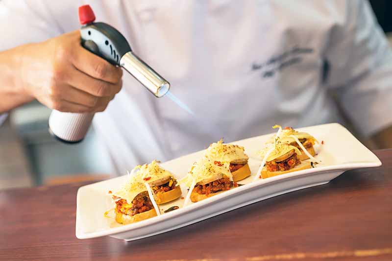 Image of a chef browning the cheese on top of appetizers on a white plate.