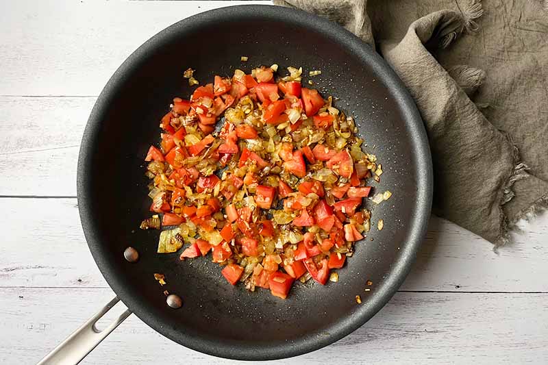 Horizontal image of a caramelized diced onion, minced garlic, and diced tomato mixture in a skillet next to a tan napkin.
