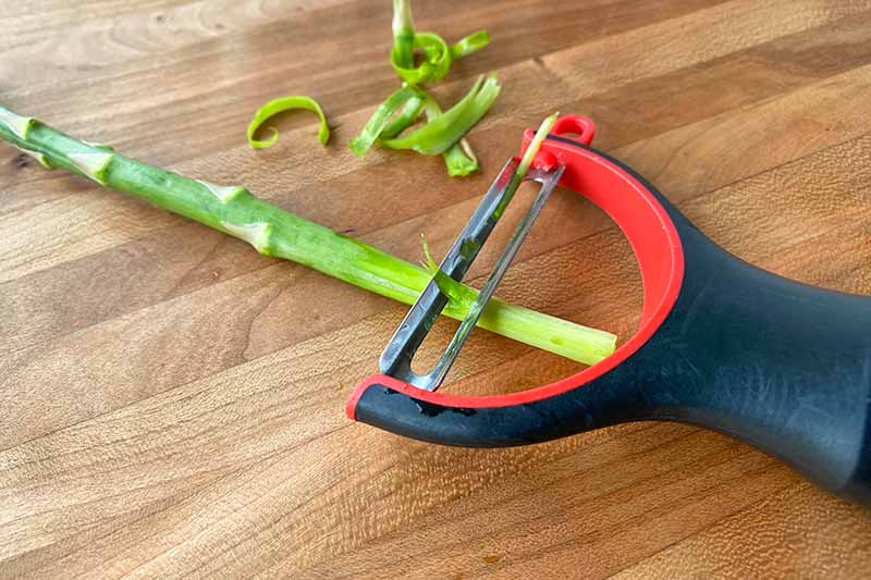 Horizontal image of peeling the outer layers of a single green vegetable using a peeler on top of a wooden cutting board.