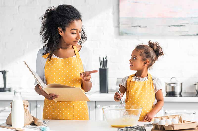 Horizontal image of a woman reading a cookbook to a child while baking. 