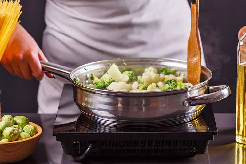 Horizontal image of a cook preparing cauliflower and broccoli on the stovetop.