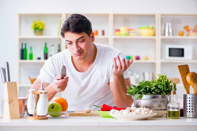 Horizontal image of a confused man holding a chef's knife next to ingredients on a table.
