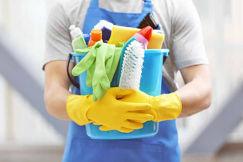 Horizontal image of a man wearing yellow dish gloves holding a bucket with assorted supplies.