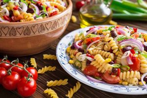 Horizontal image of a large bowl and a plate of a rotini and mixed vegetable and bean recipe next to a wooden spoon and cherry tomatoes.