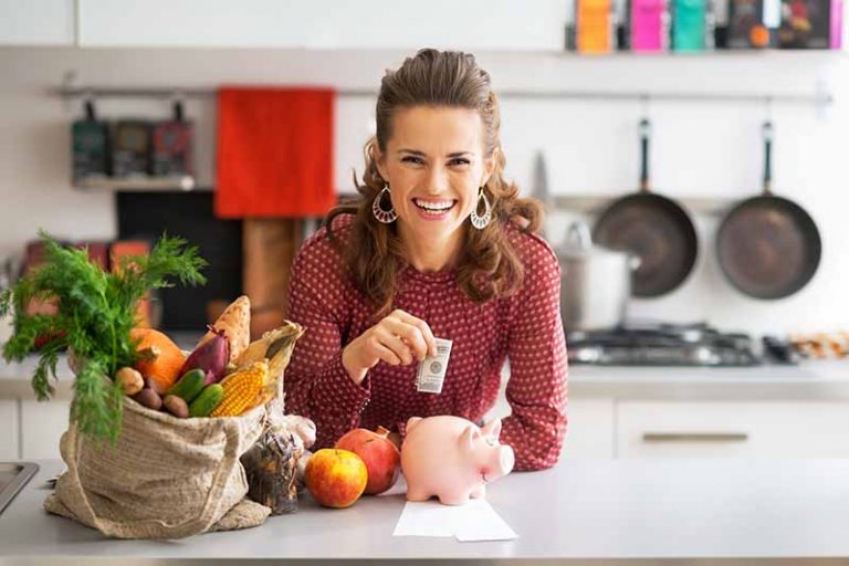 Horizontal image of a woman saving money in a piggy bank on a countertop next to groceries in a bag.