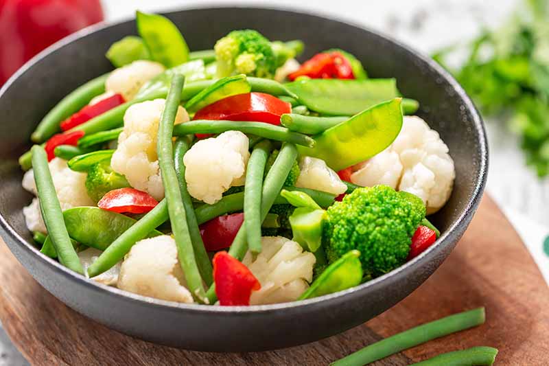 Horizontal image of a large bowl filled with cauliflower, beans, peas, and peppers.