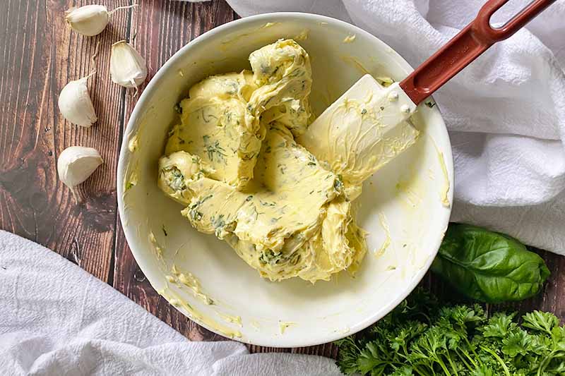 Horizontal image of a light yellow thick and creamy mixture with finely chopped greens stirred with a spatula in a white bowl.