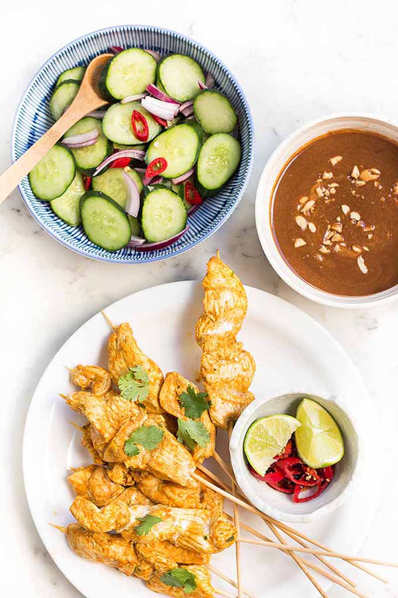 Vertical image of chicken satay on a white plate next to a bowl of cucumber salad and a bowl of peanut butter dipping sauce.