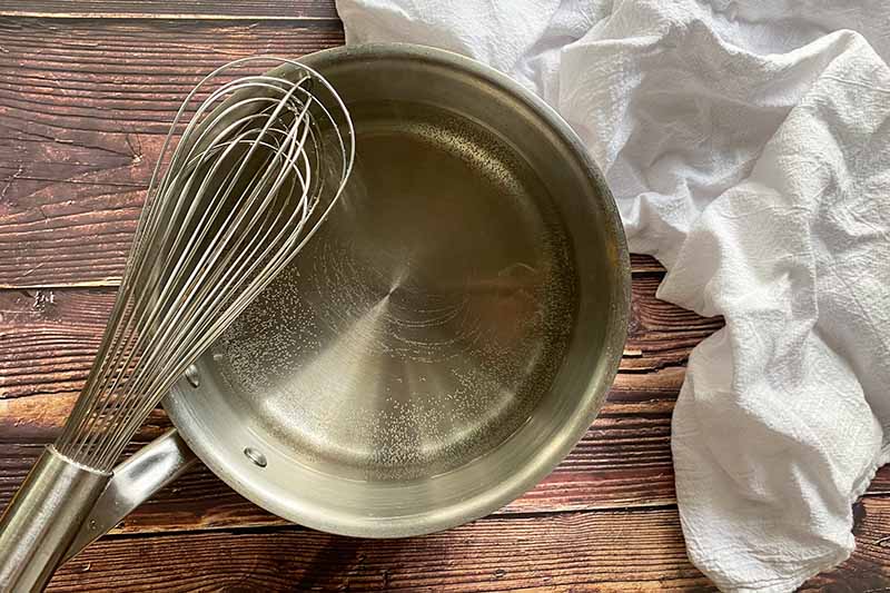 Horizontal image of a hot liquid in a pot next to a kitchen whisk and white towel.
