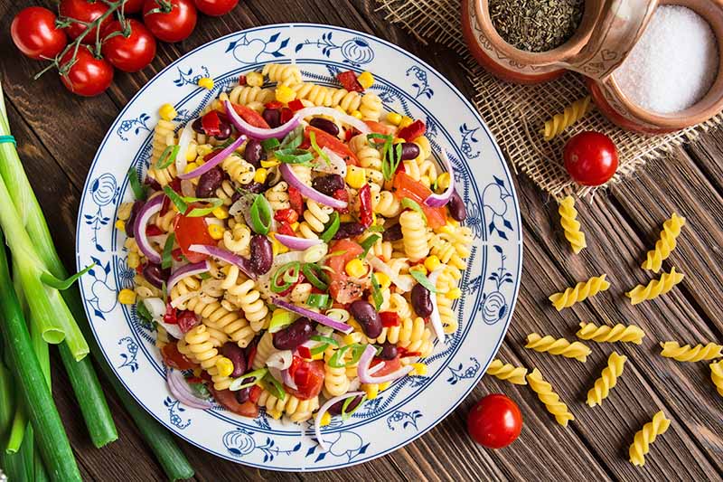 Horizontal top-down image of a plateful of a rotini and mixed vegetable and bean recipe on a wooden table next to cherry tomatoes.
