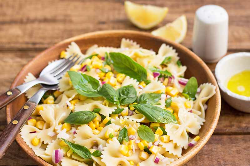 Horizontal image of farfalle mixed with red onions, corn, and basil in a wooden bowl next to a salt shaker, bowl of dressing, lemon wedges, and a metal fork.
