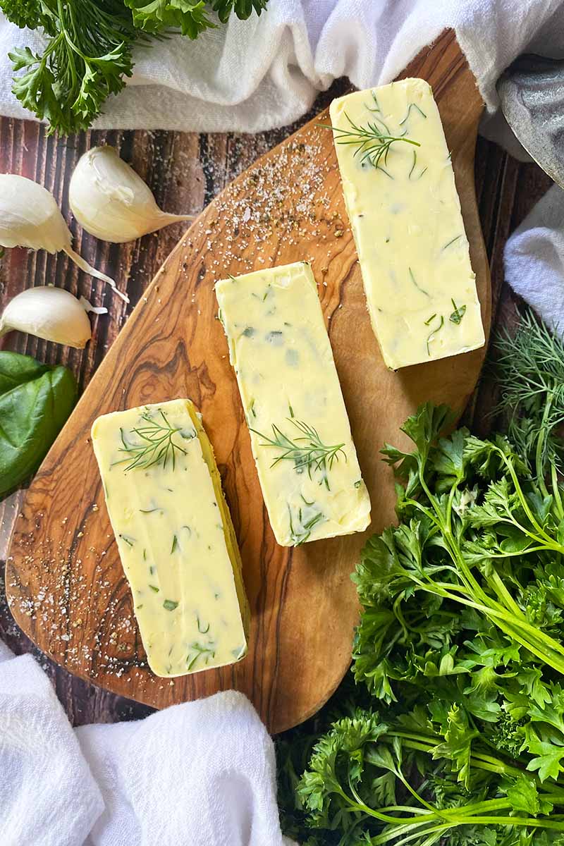 Vertical top-down image of three sticks of a light yellow mixture speckled with finely chopped greens on a wooden cutting board.