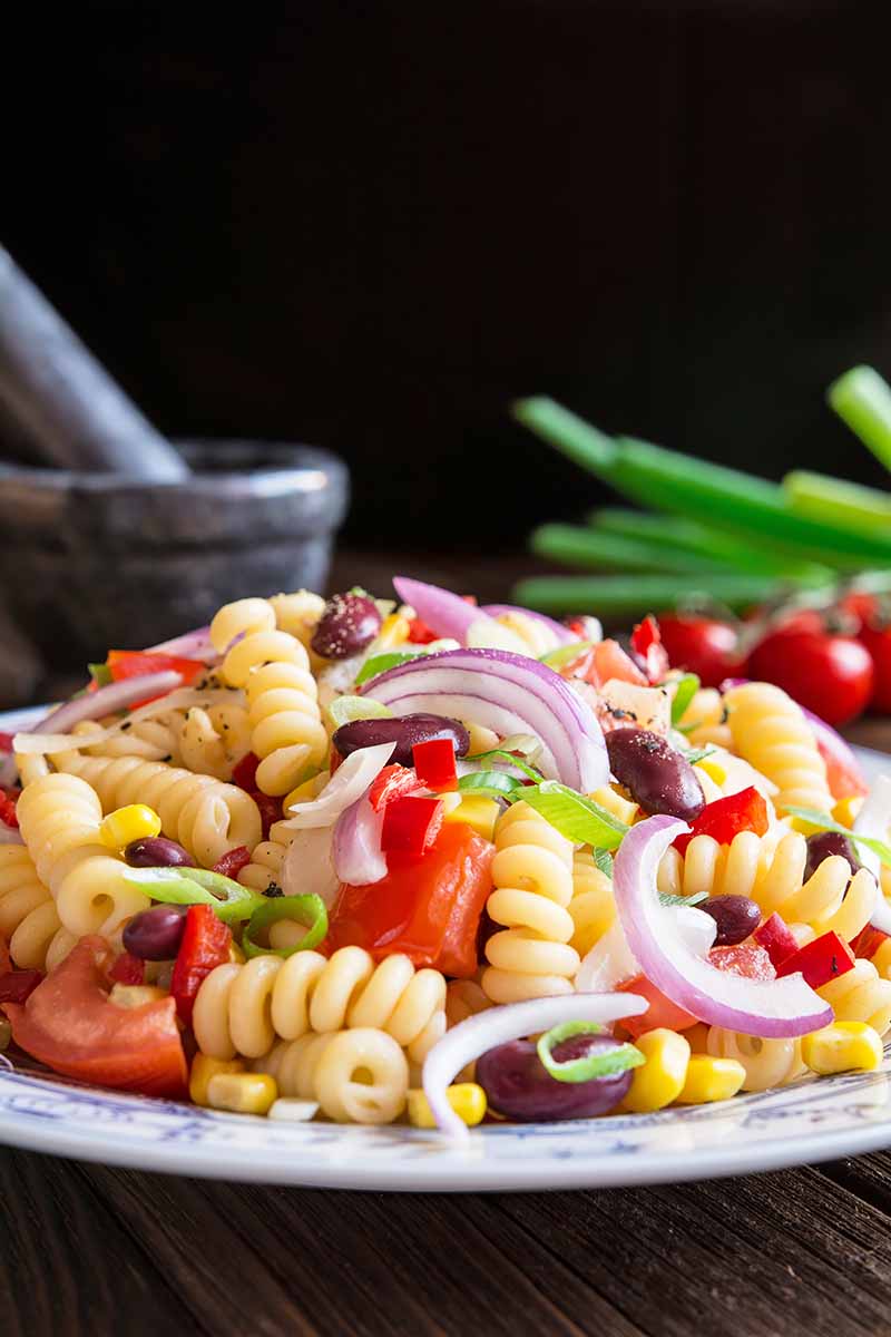 Vertical image of a plateful of a pile of rotini with assorted vegetables and seasonings on a dark surface with a dark background.
