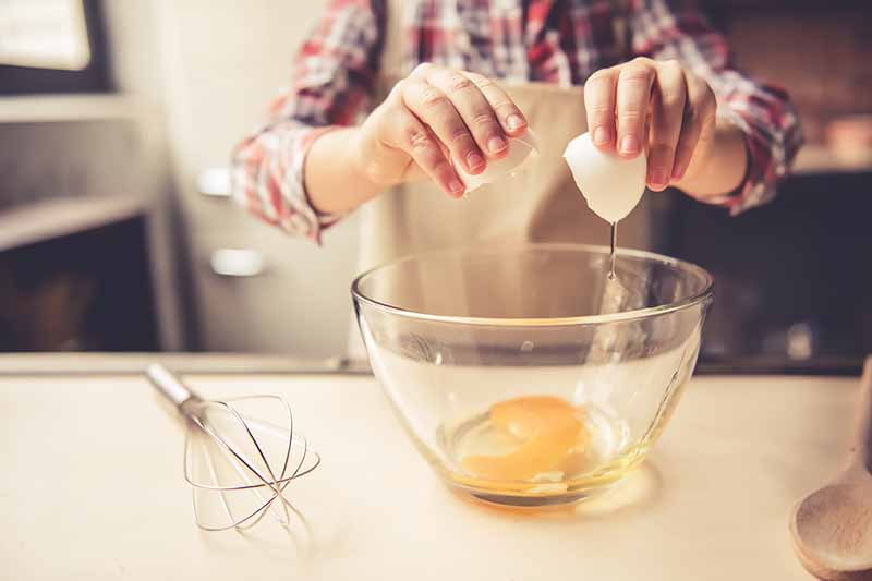 Horizontal image of a girl cracking eggs in a bowl at a table next to a whisk.