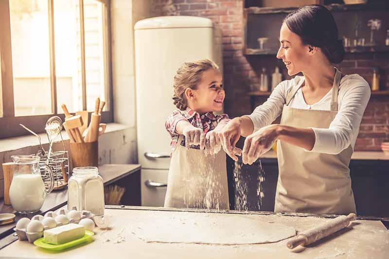 Horizontal image of a mother and daughter spreading flour on a pastry at a wooden table.