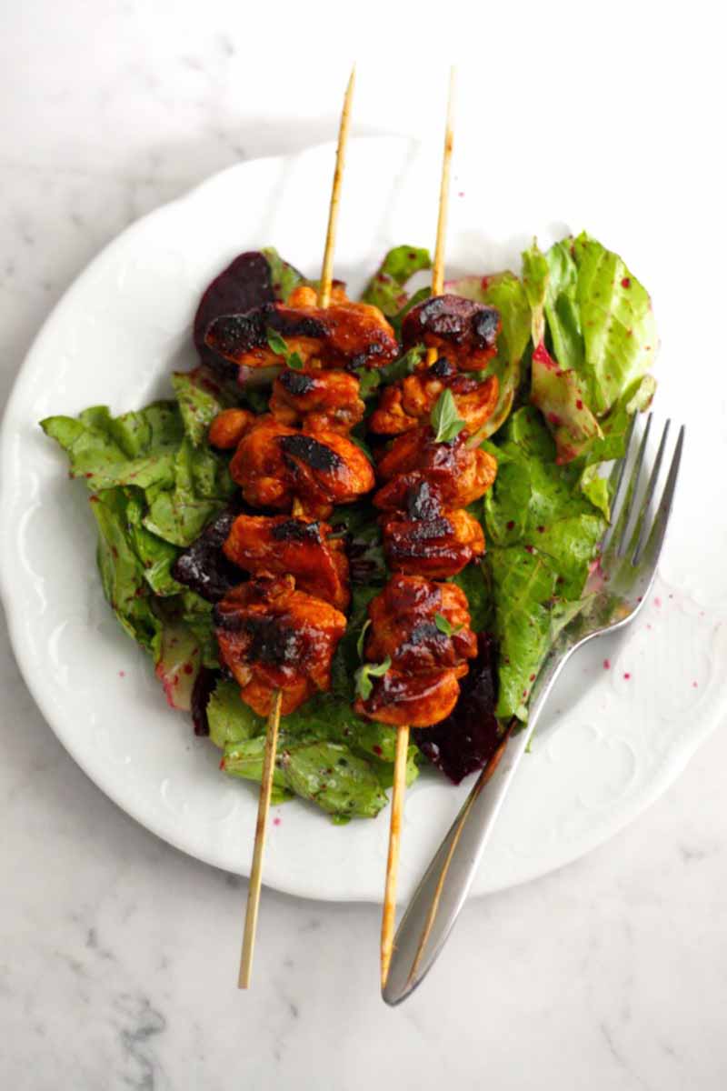 Vertical image of sambal chicken sticks over a bed of salad greens on a white plate.