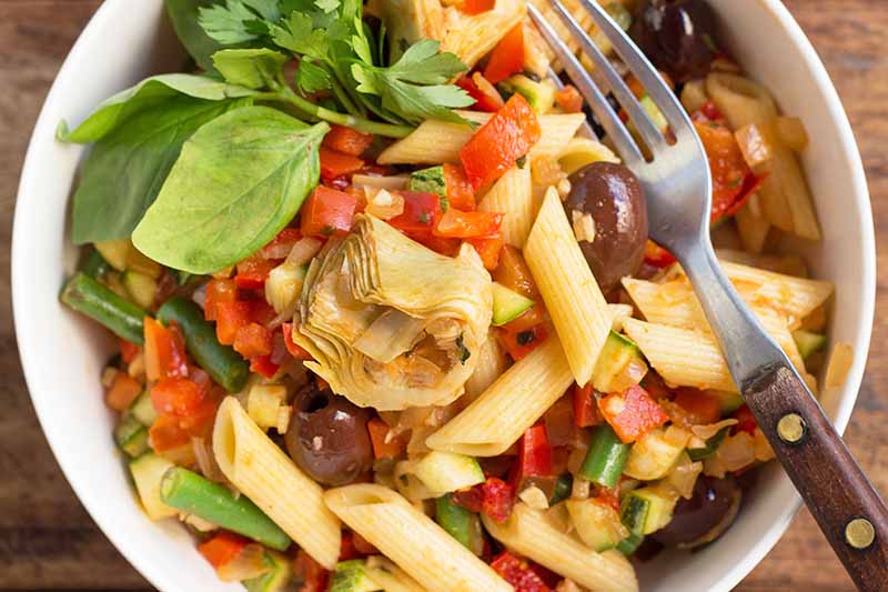 Horizontal image of penne mixed with olives, herbs, and tomatoes next to a metal fork in bowl.