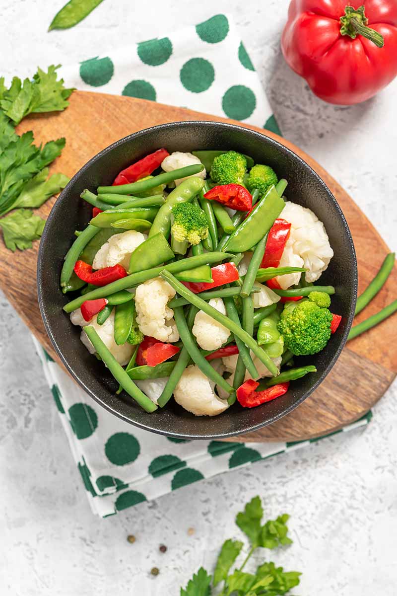 Vertical top-down image of a large bowl filled with cauliflower, broccoli, beans, peas, and bell peppers on a wooden board with a napkin.