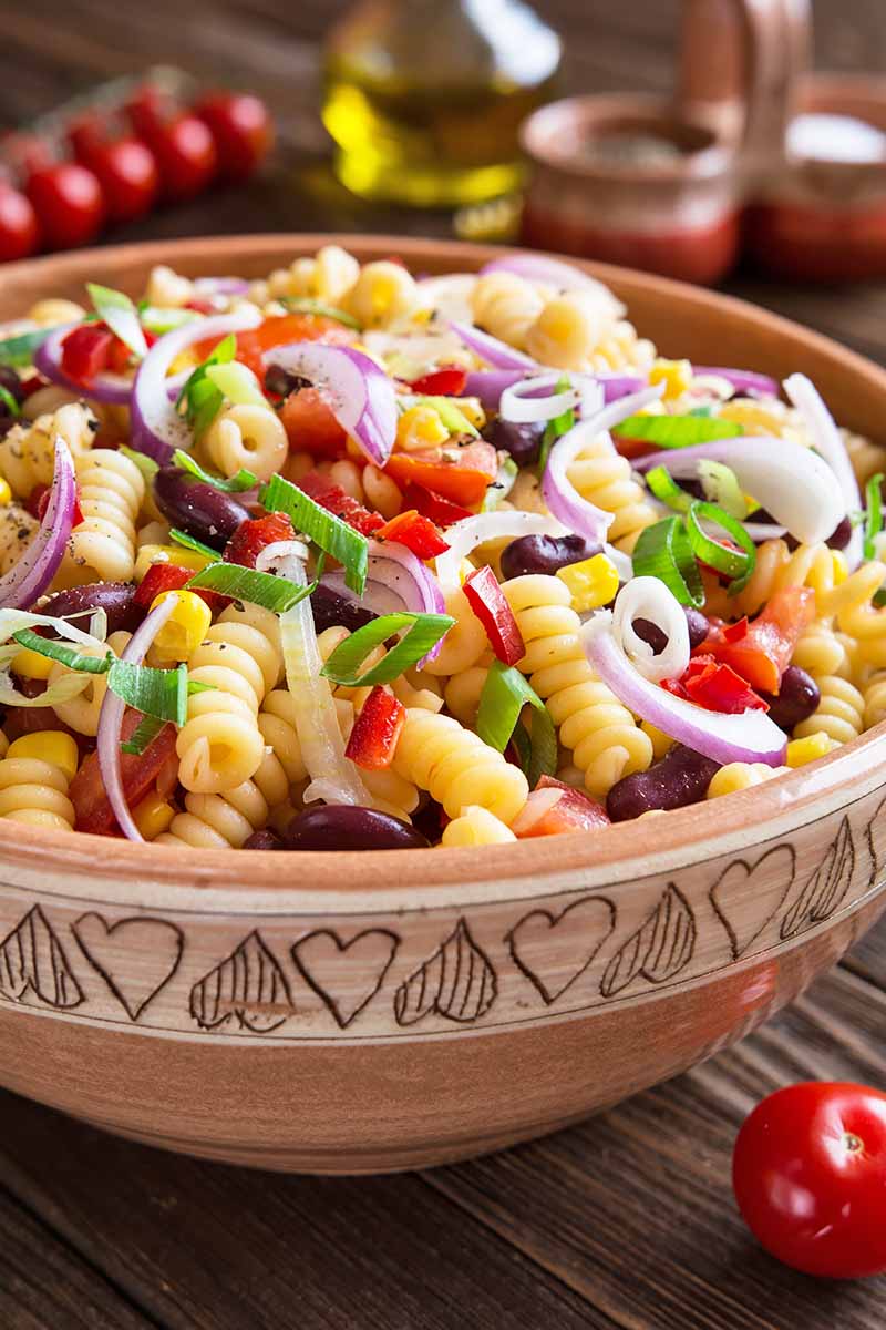Vertical image of a large bowlful of rotini mixed with assorted fresh vegetables, herbs, and beans next to tomatoes.