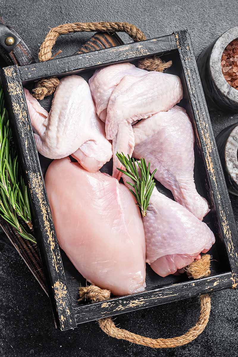 Vertical top-down image of raw cuts of poultry in a small tray with fresh herbs on top.