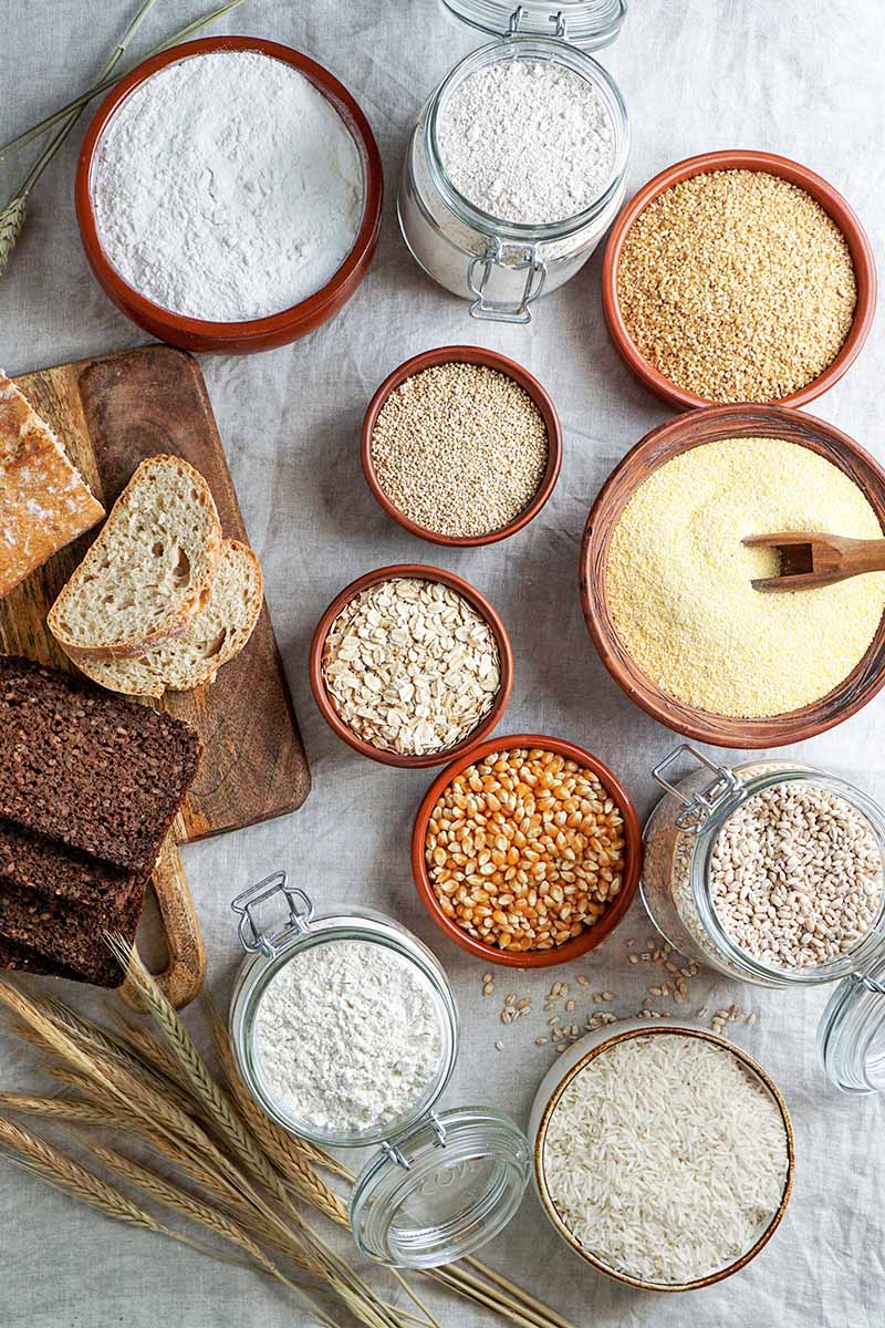 Vertical image of assorted ground and whole cereals on a table lined with a linen cloth next to bread slices.