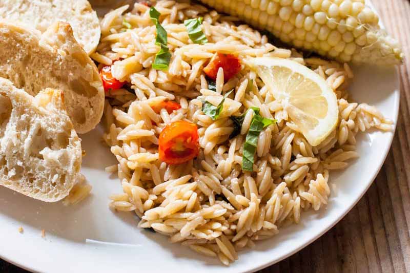 Horizontal image of orzo mixed with tomatoes and herbs on a plate next to bread, corn, and a lemon wedge.