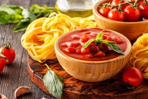 Horizontal image of a wooden bowl filled with a chunky tomato mixture next to fettuccine and fresh herbs.