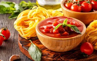 Horizontal image of a wooden bowl filled with a chunky tomato mixture next to fettuccine and fresh herbs.