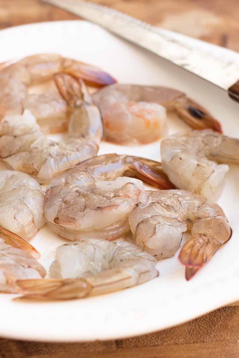 Vertical close-up image of peeled small raw crustaceans on a white plate next to a knife.