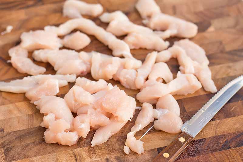 Horizontal image of slicing raw poultry in strips on a wooden cutting board.
