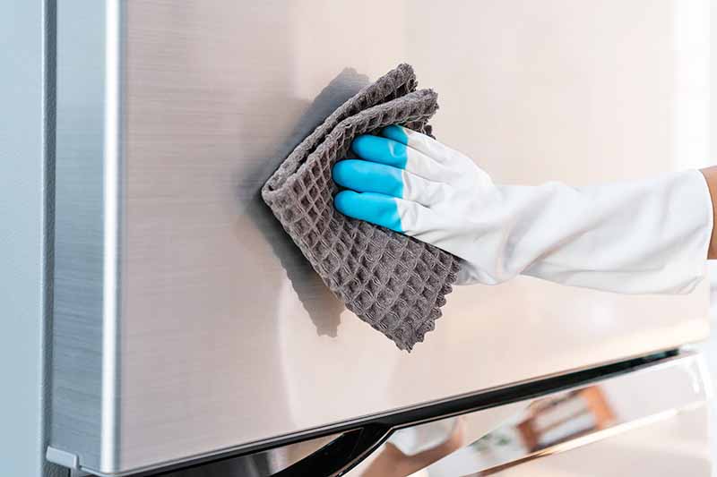 Horizontal image of a gloved hand wiping down the front of a fridge.