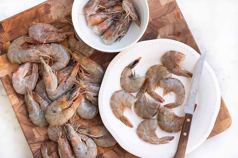 Horizontal top-down image of prepping raw prawns on a white plate and on a wooden cutting board.