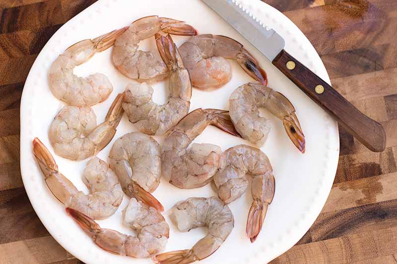 Horizontal image of peeled and deveined raw small crustaceans on a white plate next to a knife.
