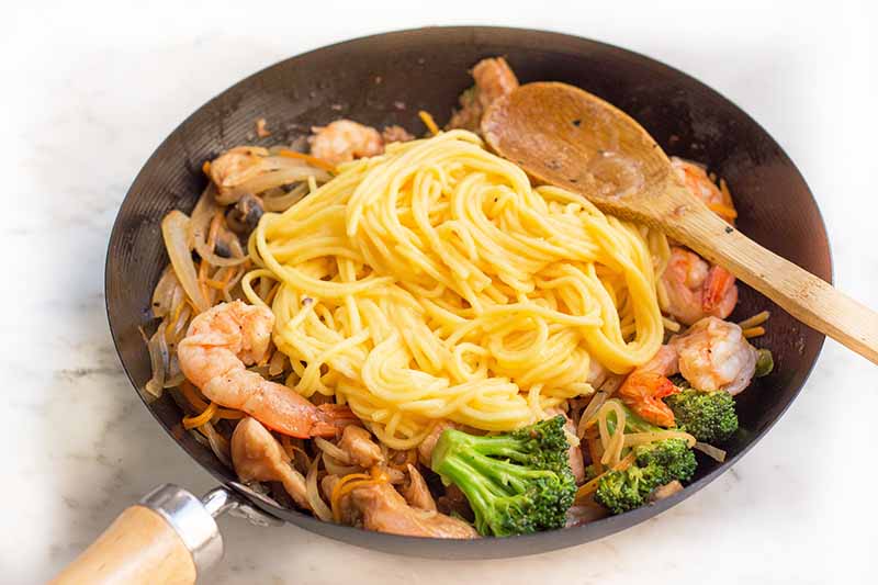 Horizontal image of a pile of egg noodles on a pile of mixed veggies in a pan stirred by a spoon.