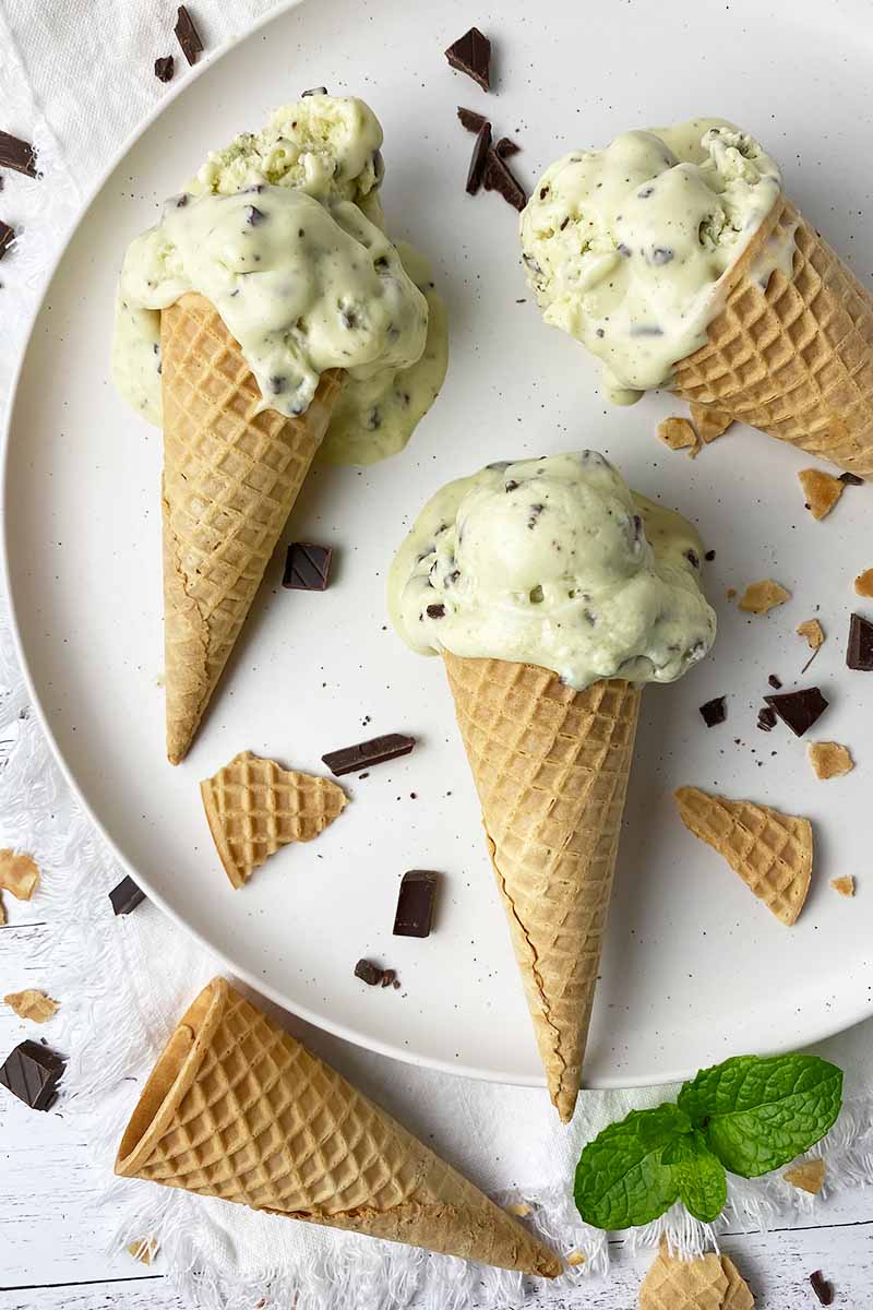 Vertical top-down image of three sugar waffle cones filled with light green ice cream next to chopped candies and fresh herbs.
