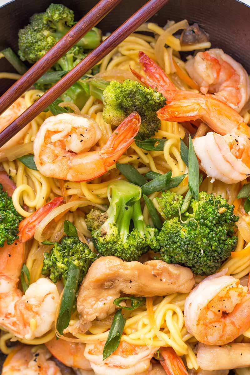 Vertical close-up image of shrimp, broccoli, chicken strips, and noodles.