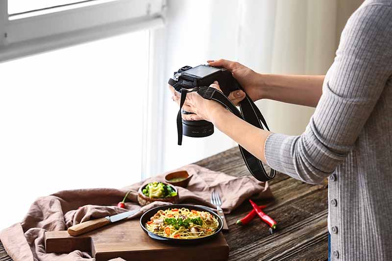 Horizontal image of a woman taking a picture of a food scene in front of a window.