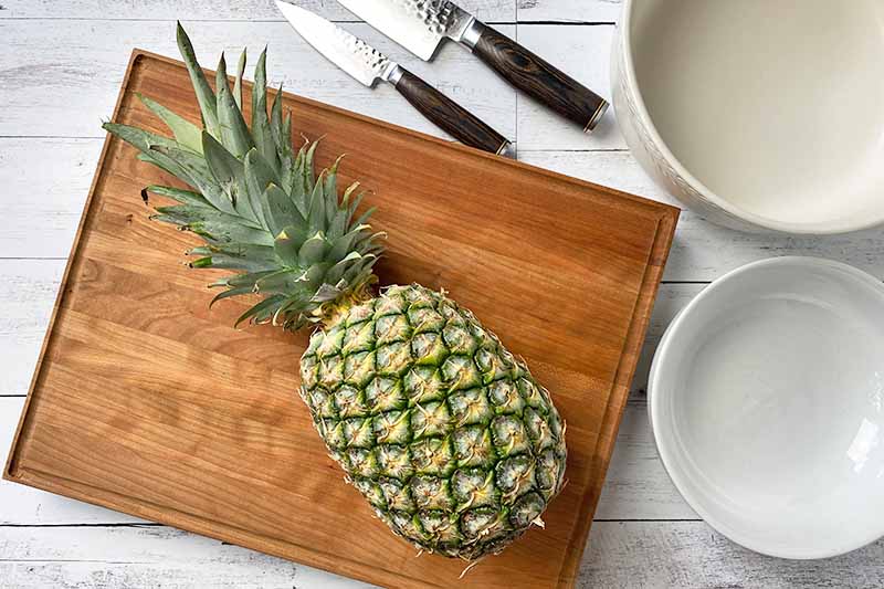Horizontal image of a whole fruit on a wooden cutting board next to knives and empty bowls.