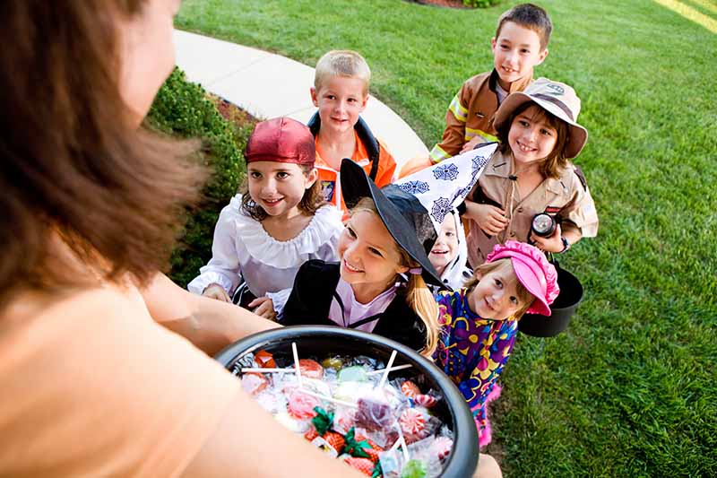 Horizontal image of a woman handing out candy to a group of children during Halloween.