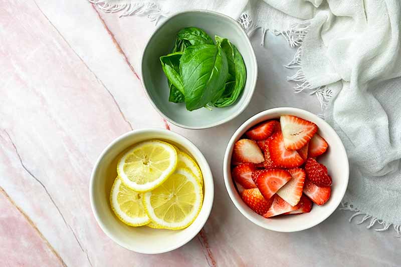 Horizontal image of bowls of sliced lemon, strawberry, and whole basil leaves.