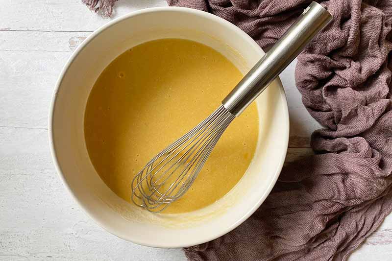 Horizontal image of a yellow liquid in a bowl with a whisk.