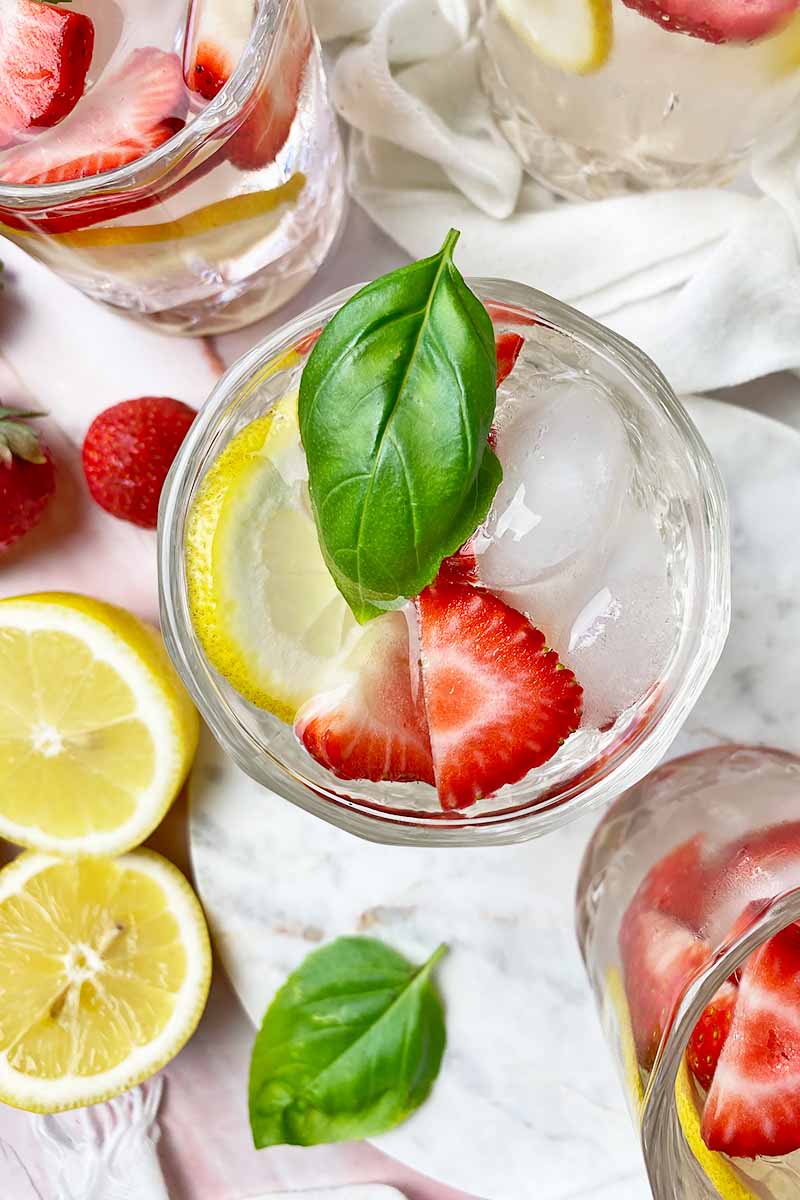 Vertical top-down image of glasses filled with a clear liquid mixed with slices of strawberry and lemon and garnished with basil.