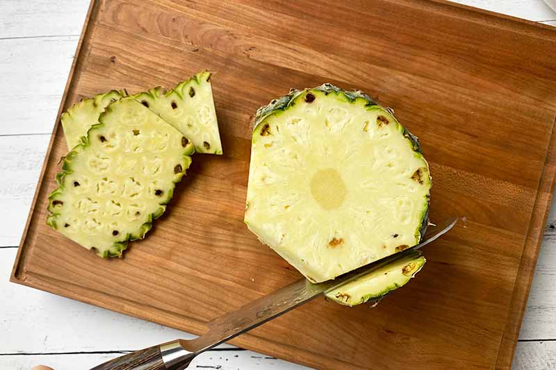 Horizontal image of slicing off the skin of a pineapple on a wooden cutting board.